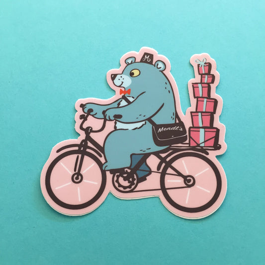 die cut sticker of a blue bear riding a bike on a pastel pink background